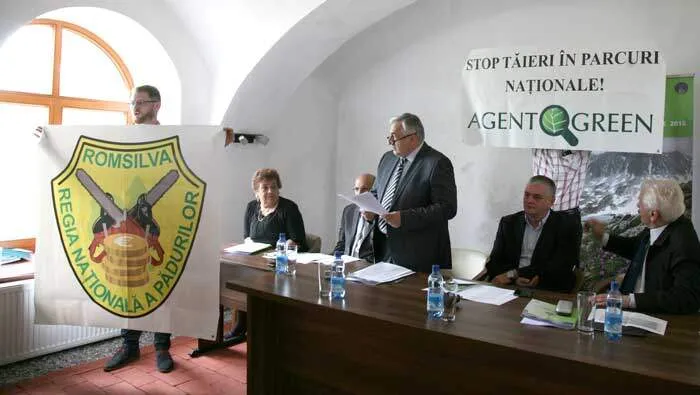 Participanți la o întâlnire, lângă însemnele Romsilva și pancarta Agent Green „Stop tăieri în parcuri naționale!”