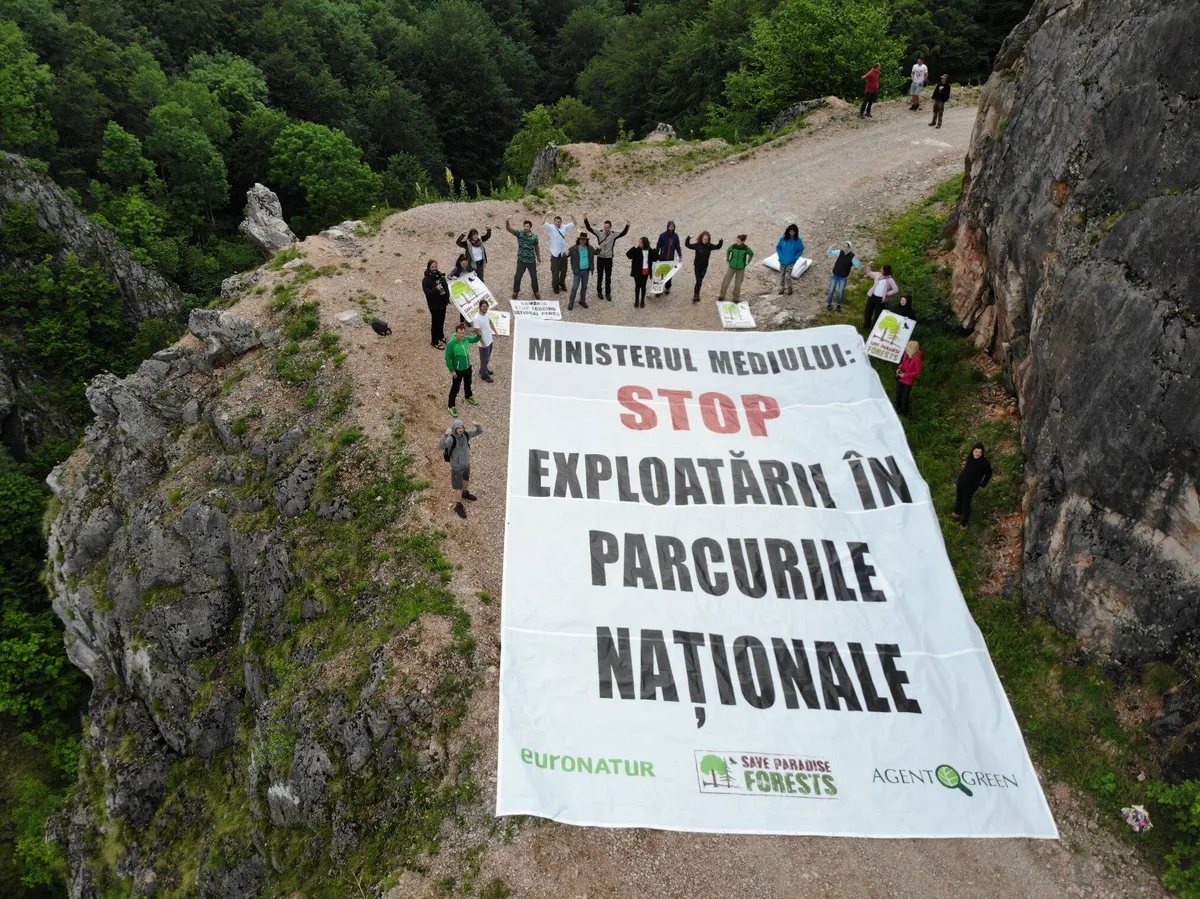 Manifestanți ținând pe un drum montan o pancartă cu mesajul „Ministerul Mediului! STOP EXPLOATĂRI ÎN PARCURILE NAȚIONALE”.