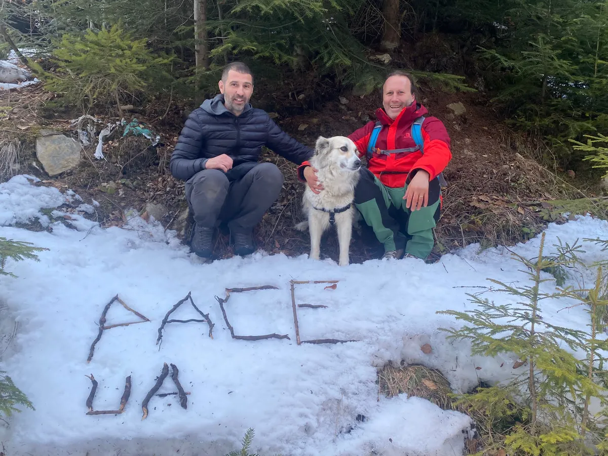 Doi bărbați și un câine într-o pădure, lângă cuvântul „PACE” scris pe zăpadă.