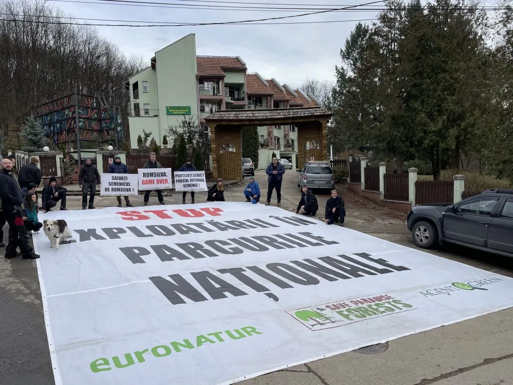 Banner împotriva exploatării în parcurile naționale întins în fața unei clădiri, alături de protestatari cu pancarte.