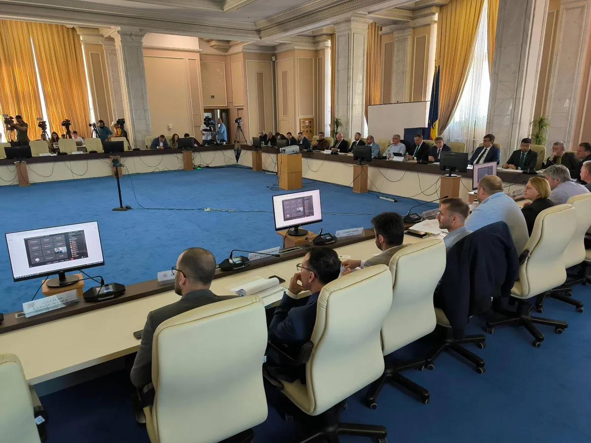 Participanți așezați în jurul unei mese de conferință, într-o sală cu draperii galbene.